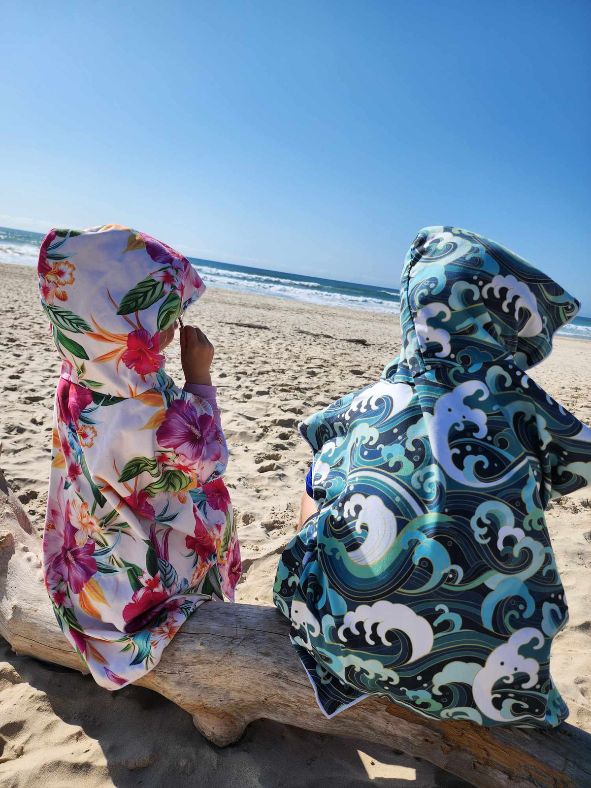 Two little ones in their sand less hooded beach towel one in a Island Princess the other a Wipeout both are sitting on a log overlooking the beach