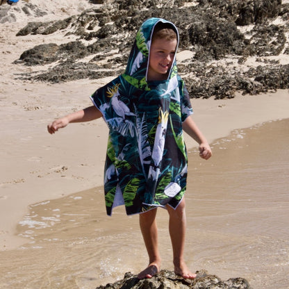 Boy (ages 6-12) wearing a blue hooded beach hoodie with a Bird of Paradise design featuring a yellow crested white cockatoo and palm leaves. He is standing on a rock at Currumbin Beach, Queensland, with a big smile on his face and arms outstretched, looking out at the ocean.
