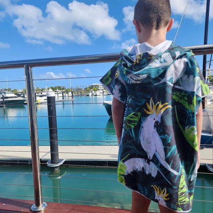 Boy (ages 6-12) wearing a blue hooded beach hoodie with a Bird of Paradise design featuring a yellow crested white cockatoo and palm leaves. He is standing on a pontoon at Hamilton Island, Queensland, looking out over moored boats and sailing vessels in the Whitsunday Islands.