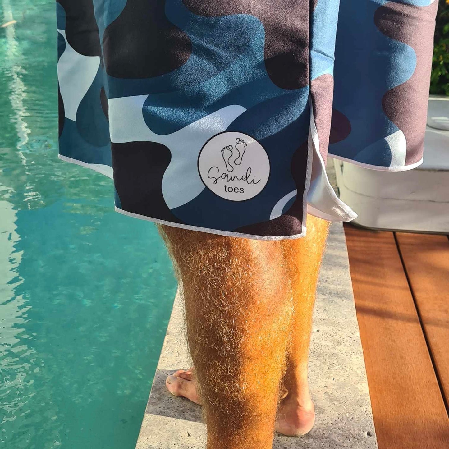 Camouflage Sand-Free Hoodie (One Size Fits All): Poolside Relaxation in Style. [Adult male in camouflage hoodie enjoys a moment beside a private swimming pool.] Shows Sandi Toes Logo.
