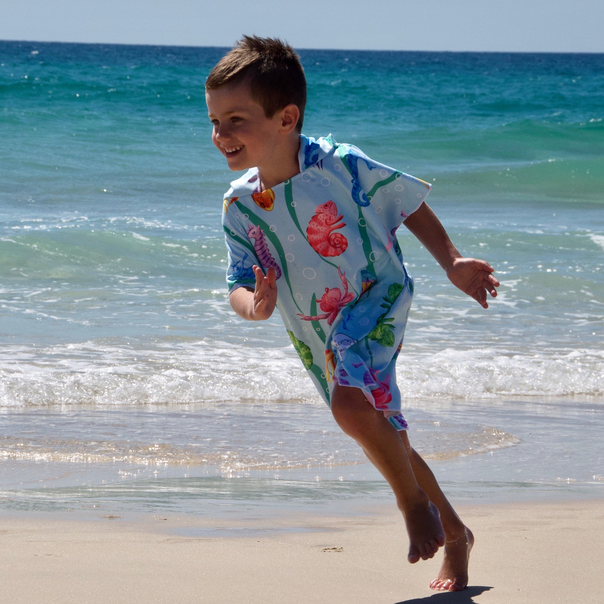 Boy (ages 6-12) wearing a [hoodie color] Earth & Sea hooded beach hoodie. The design features a vibrant underwater scene teeming with sea creatures (colorful fish, chameleon, crabs, seahorse, jellyfish). He is running fast on the beach with waves visible in the background.