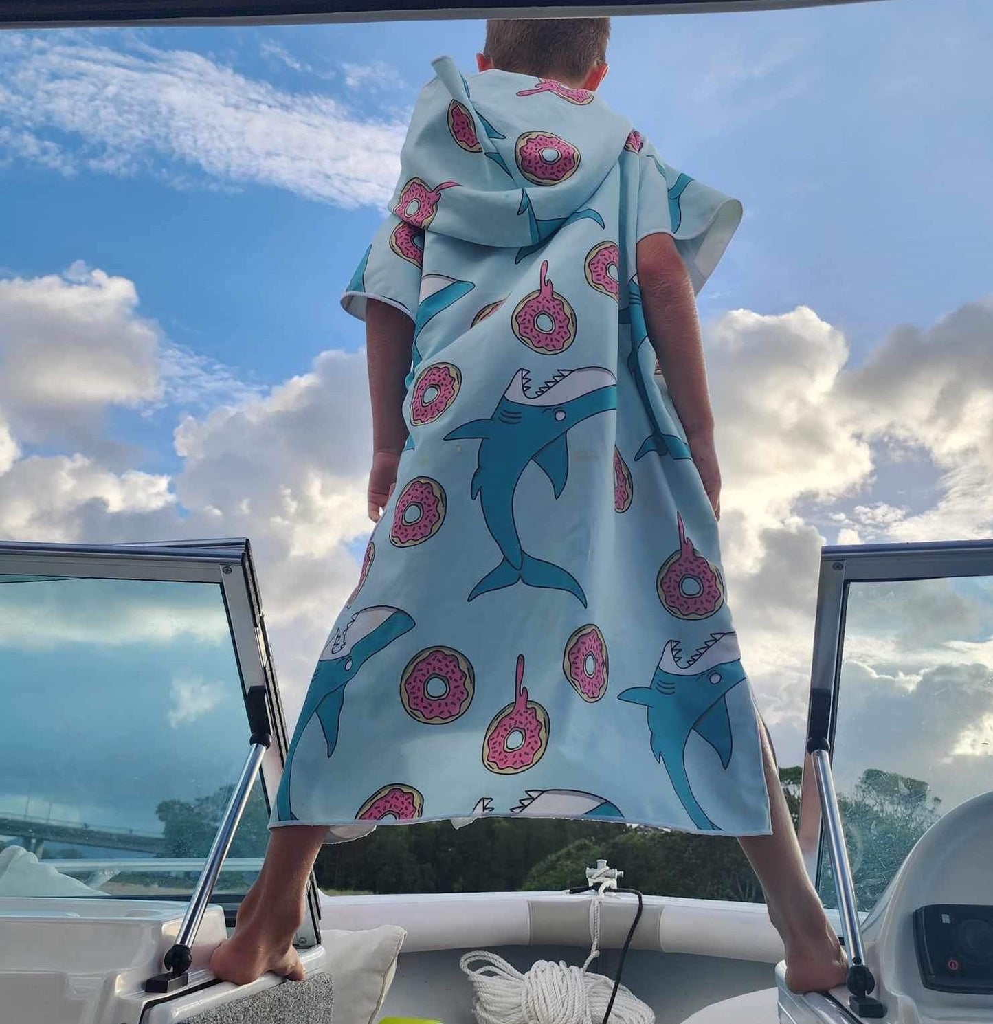 Boy (ages 6-12) wearing a Feeding Frenzy hooded beach towel. The design features a playful scene with colourful fish swimming around dripping donuts. He is standing excitedly on his dad's boat at Currumbin Beach, Queensland, ready for a fun day at beach.