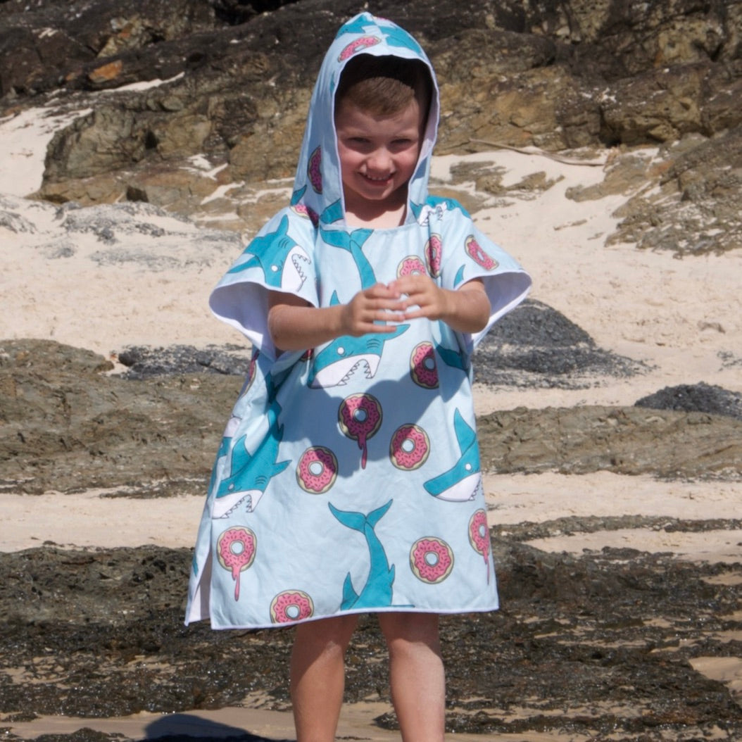 Toddler boy (ages 1-3) wearing a Feeding Frenzy hooded beach towel (playful scene with colourful sharks and dripping donuts) with the hood pulled up for sun protection. He is facing the camera and clapping his hands in delight while standing on the beach at Currumbin, Queensland.