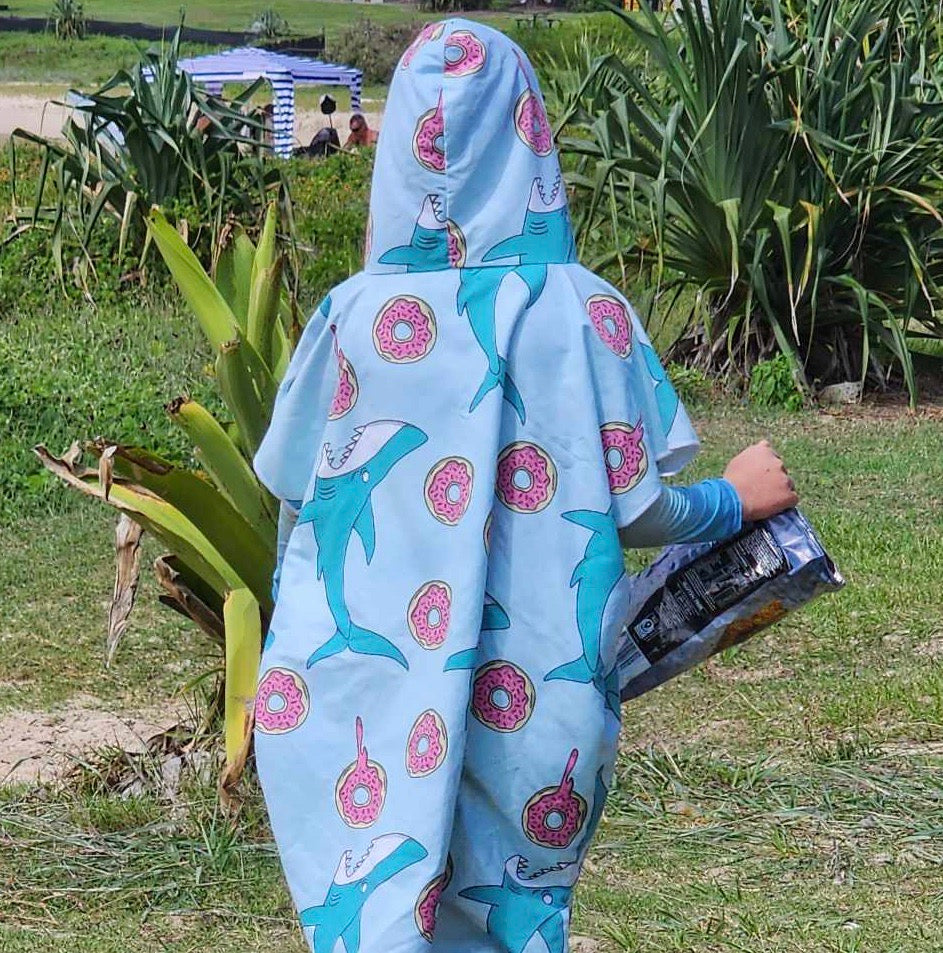 Boy (ages 6-12) wearing a Feeding Frenzy hooded beach towel (playful scene with colourful fish and dripping donuts) with the hood pulled up over his head. He is standing on the beach at Hastings Point, New South Wales, with his back to the camera.
