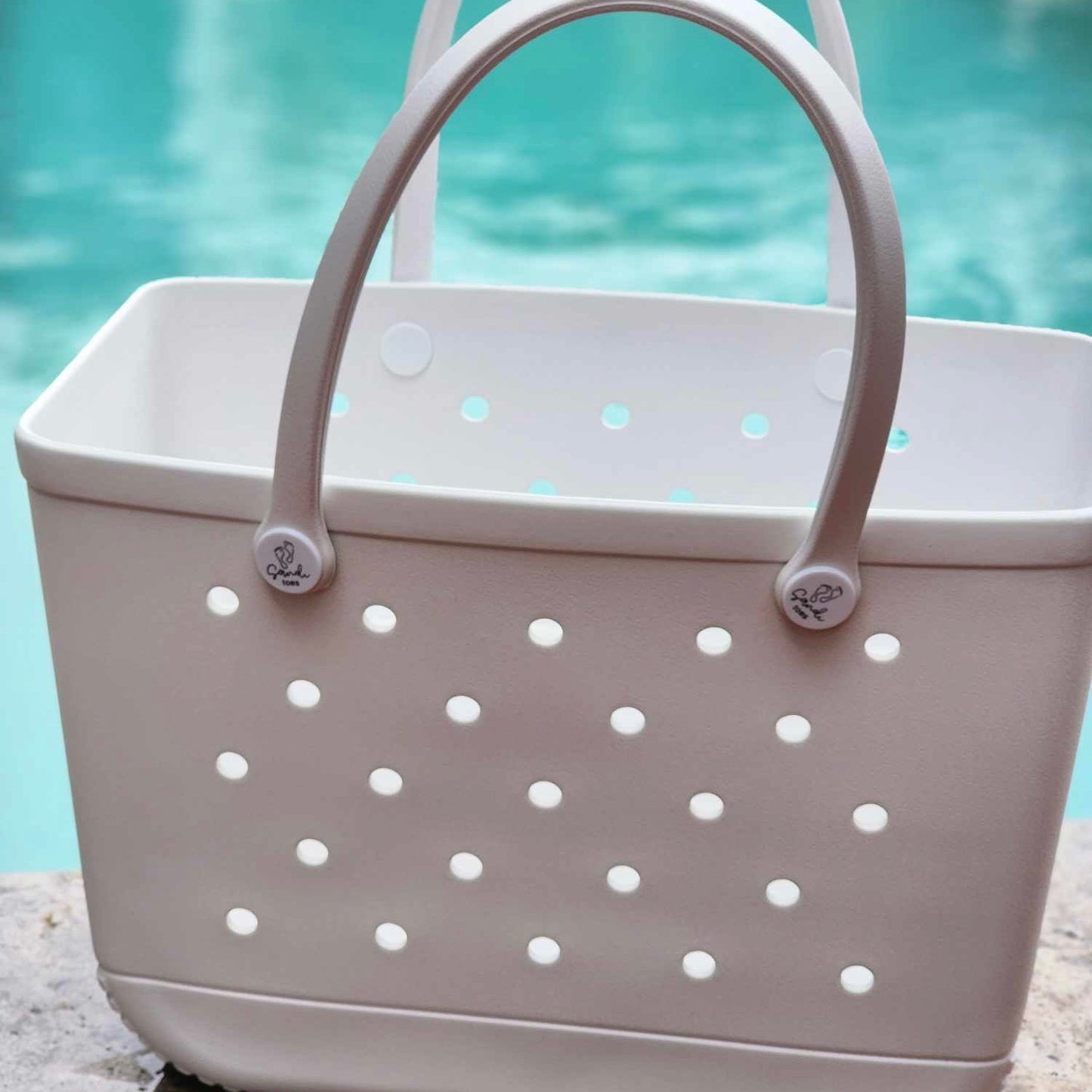 White Haven Silicone Bag (Sandi Toes): Poolside Essentials in Style. [White silicone bag with the Sandi Toes logo rests beside a sparkling pool, perfect for keeping your swim essentials organised.]