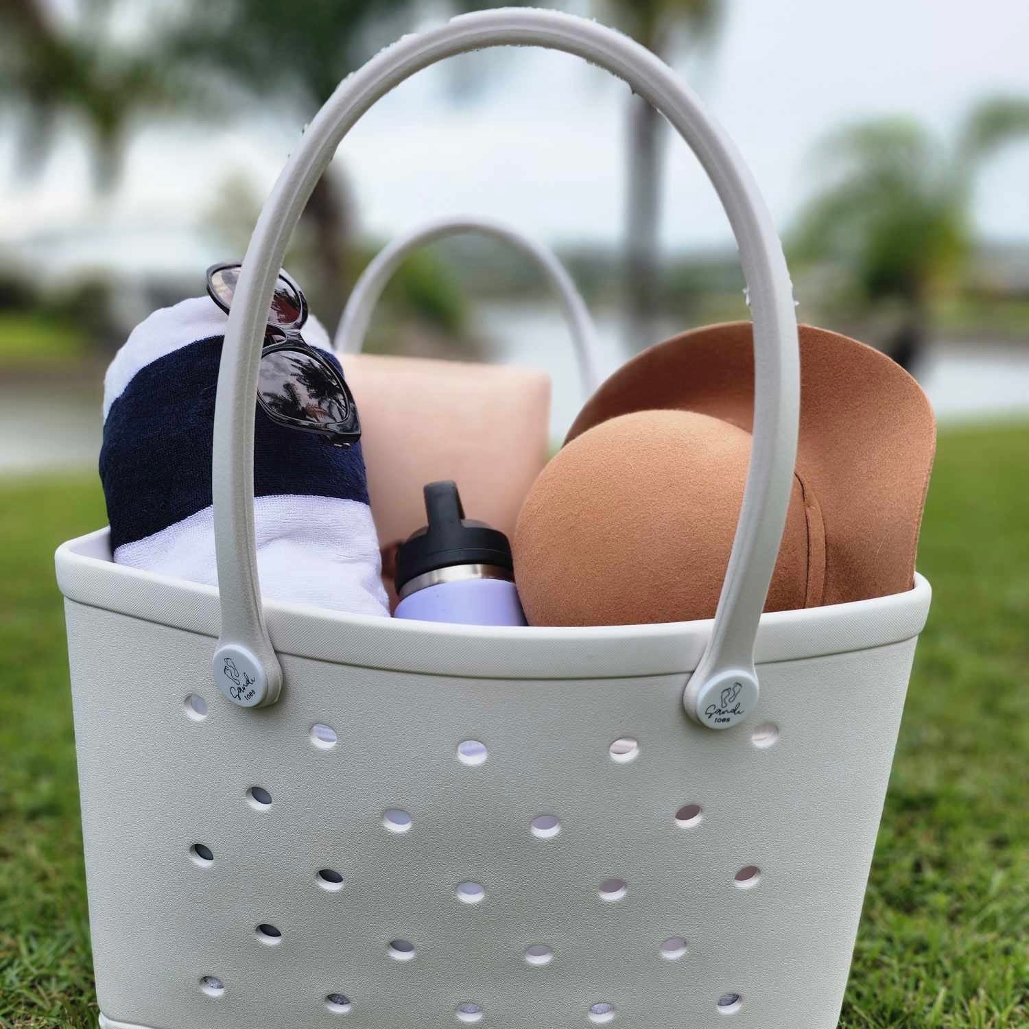 White Haven Silicone Bag (Sandi Toes): Beach Essentials in Style. [Spacious white silicone bag with the Sandi Toes logo holds sunglasses, hat, hoodie, water bottle, and phone - all set for a fun day at the beach.]