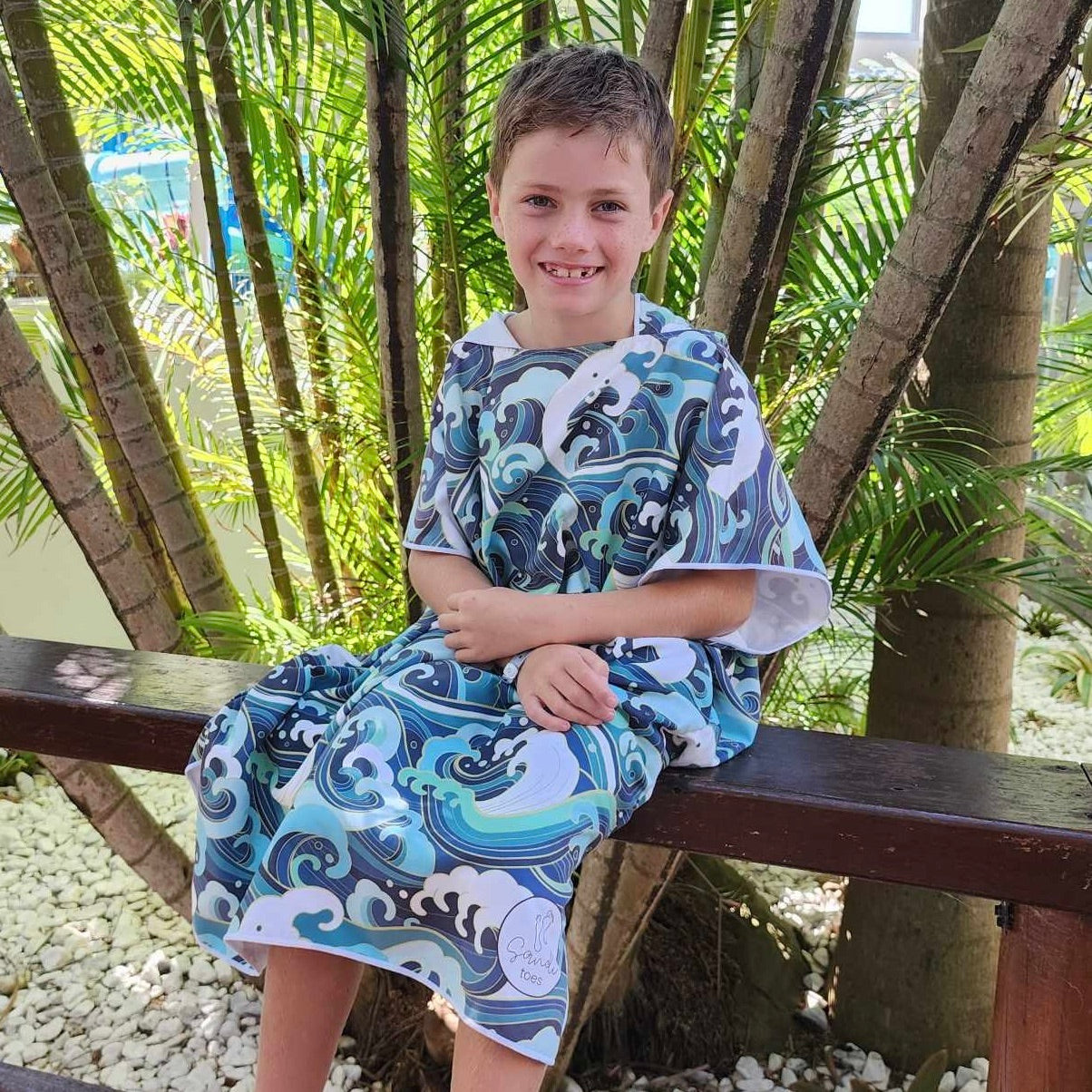 Boy (ages 6-12) sitting on a rail at Hastings Point Caravan Park. He is wearing a Wipeout hooded beach towel featuring a vibrant design inspired by the rolling waves of Currumbin Beach. The towel is sand-free, making it a convenient beach companion.