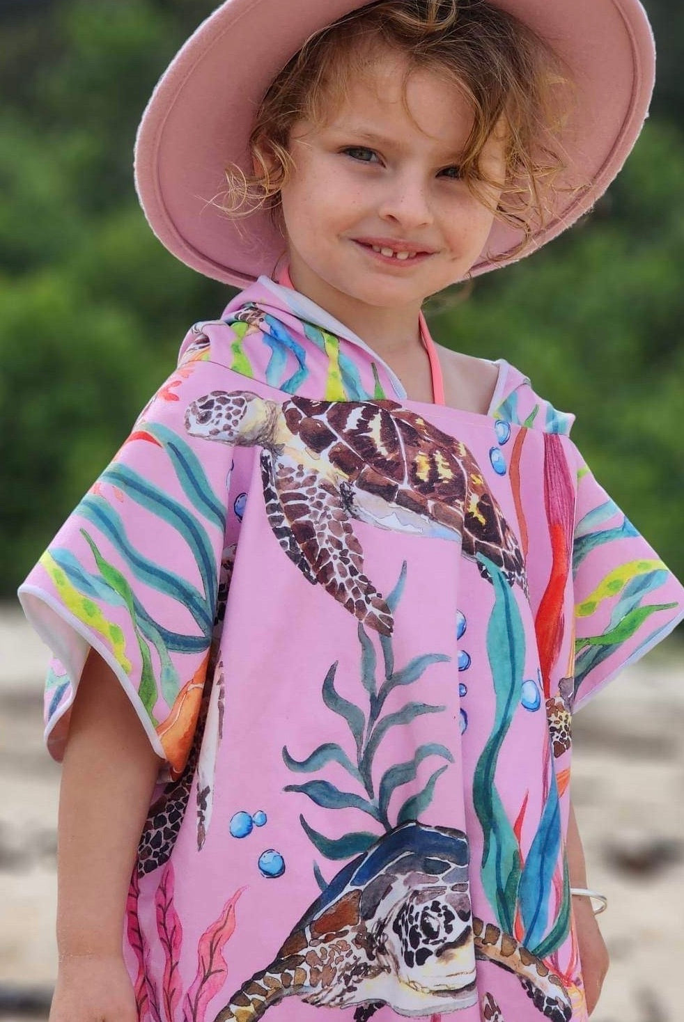 Behold the beauty of a 5-year-old girl adorned in the most popular design from Sandi Toes' sand-free hooded towels collection – 'Turtle Cove.' This captivating print features turtles gracefully swimming amidst sea grasses on a predominantly pink background. Now available in Large sizes, in addition to Small and Medium, this towel is your perfect companion for any adventure. The image also showcases the girl wearing a pink wide-brimmed hat (not included)