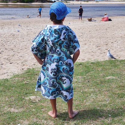 Boy (ages 6-12) wearing a Wipeout hooded beach towel and a hat. The towel features a vibrant design inspired by the rolling waves of Currumbin Beach. He is sitting on a rail at Hastings Point Caravan Park, looking out over the beach.