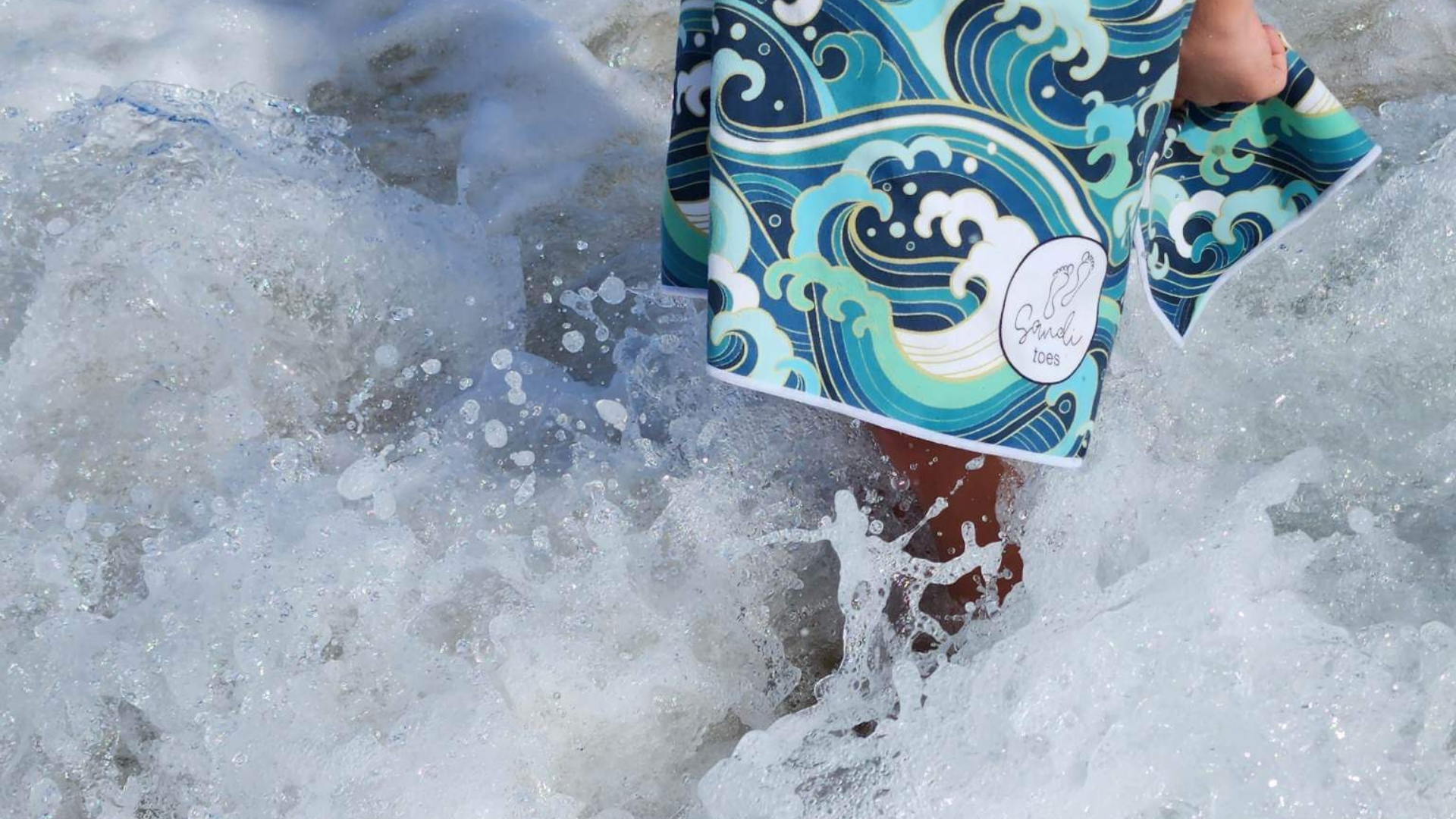 "Dive into the swirling foaming waves with Sandi Toes' 'Wipeout' Sand-Free Hooded Beach Towel! This captivating image showcases the Wipeout design – tropical waves that captivate the essence of beach adventures. Featuring the Sandi Toes logo on the towel, the foaming ocean waves swirl around a boy's foot, creating a picturesque scene. Wipeout is now available in Small, Medium, and Large sizes – your ideal companion for every adventure!