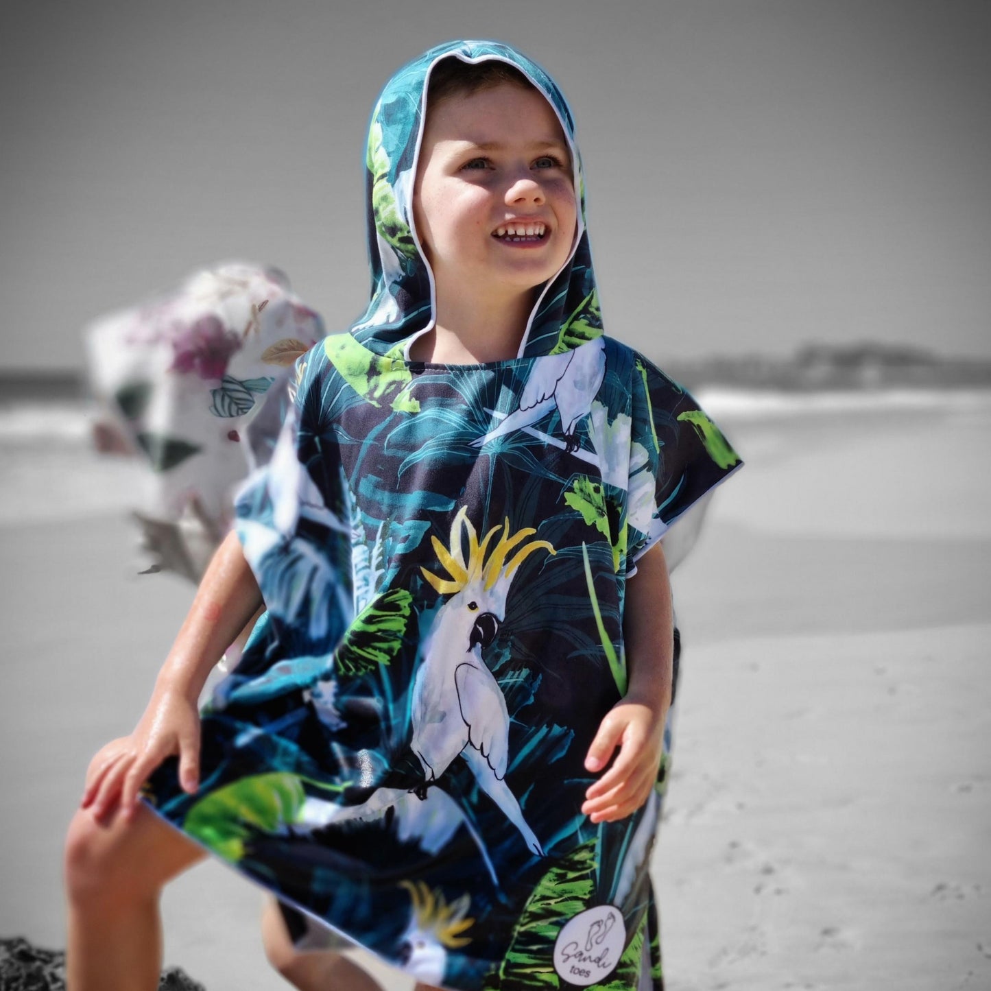 Boy (ages 6-12) wearing a blue hooded beach hoodie with a Bird of Paradise design featuring a yellow crested white cockatoo and palm leaves. He is standing at Currumbin Beach, Queensland, with one foot placed on a rock and the other in the shallow water. He has a relaxed and smile expression on his face.