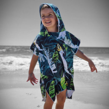 Boy (ages 6-12) wearing a blue hooded beach hoodie with a Bird of Paradise design (yellow crested white cockatoo and palm leaves) pulled up over his head. He is smiling on a beach at Currumbin, Queensland.