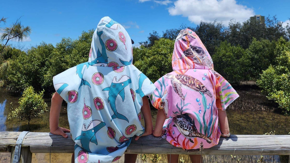 Two kids sitting on a fence wearing their sand free hoodies left is Feeding Frenzy and the right is Turtle Cove
