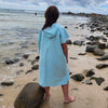 Adult model wearing a clear blue Aqua Lagoon Sand-Free Hoodie (back facing camera). Model is standing relaxed by water. Sand-free hoodie for comfortable beach adventures.