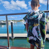 Boy (ages 6-12) wearing a blue hooded beach hoodie with a Bird of Paradise design featuring a yellow crested white cockatoo and palm leaves. He is standing on a pontoon at Hamilton Island, Queensland, looking out over moored boats and sailing vessels in the Whitsunday Islands.