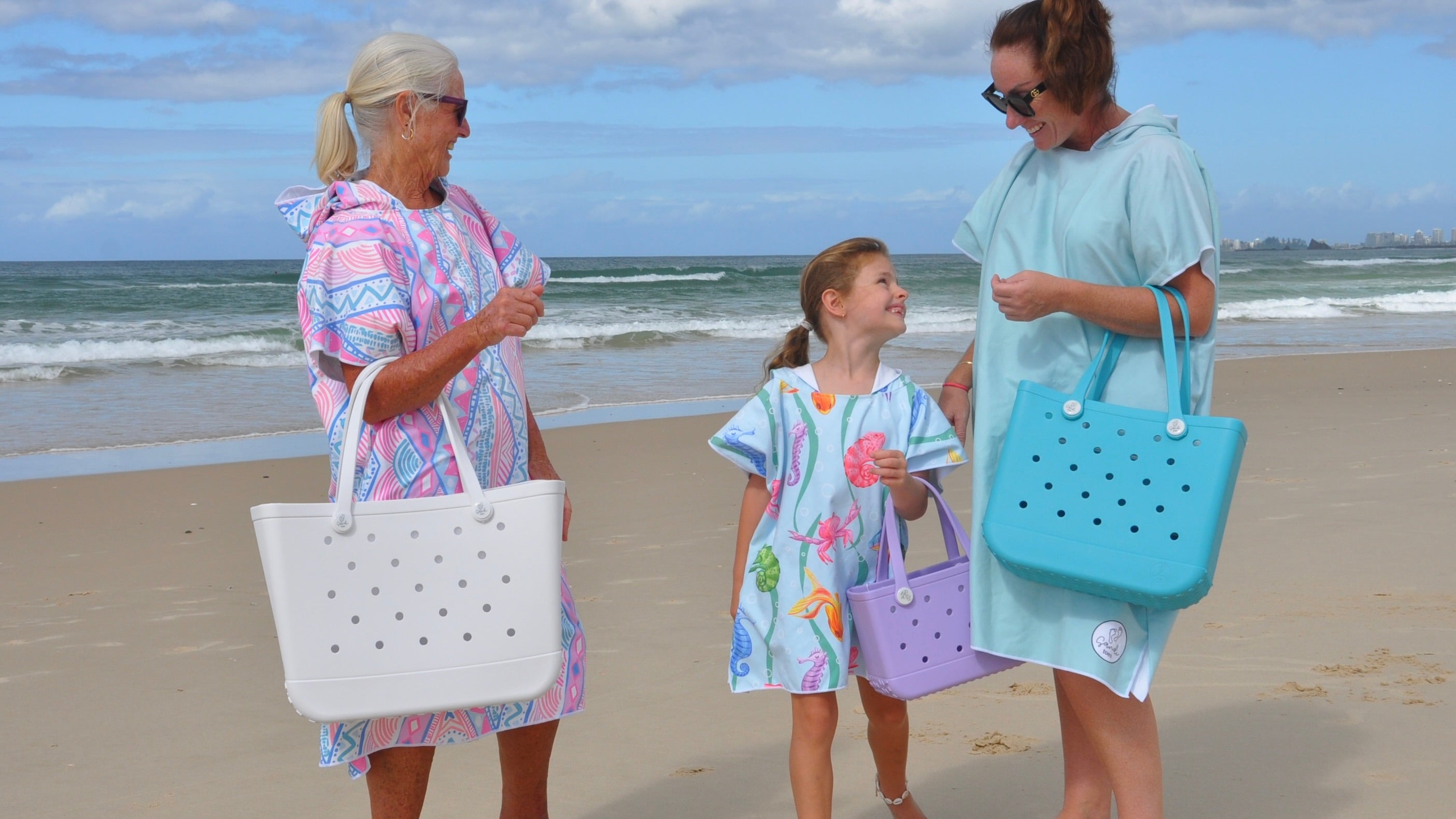 Beach image of Grand Mother Grand Daughter and Mother wearing Adult and Kids Sand Free Hoodies, on Palm Beach Queensland Australia. Xalli adult, Earth and Sea Kids and Aqua Lagoon Adult with 3 EVA silicone bags The Hauler White, The Mini Coral Cove and The Everyday Aqua Marine.