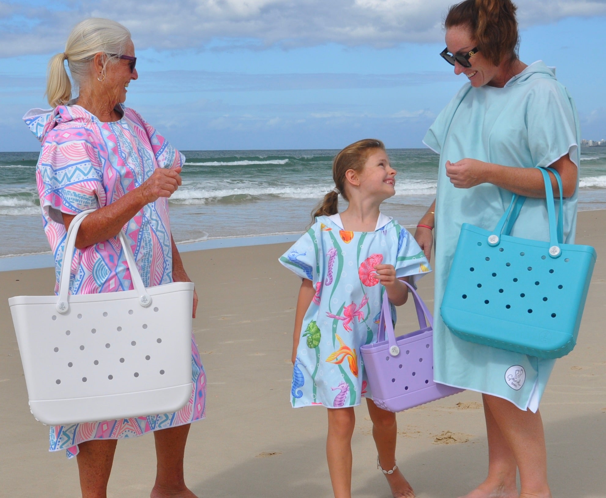 Beach image of Grand Mother Grand Daughter and Mother wearing Adult and Kids Sand Free Hoodies, on Palm Beach Queensland Australia. Xalli adult, Earth and Sea Kids and Aqua Lagoon Adult with 3 EVA silicone bags The Hauler White, The Mini Coral Cove and The Everyday Aqua Marine.
