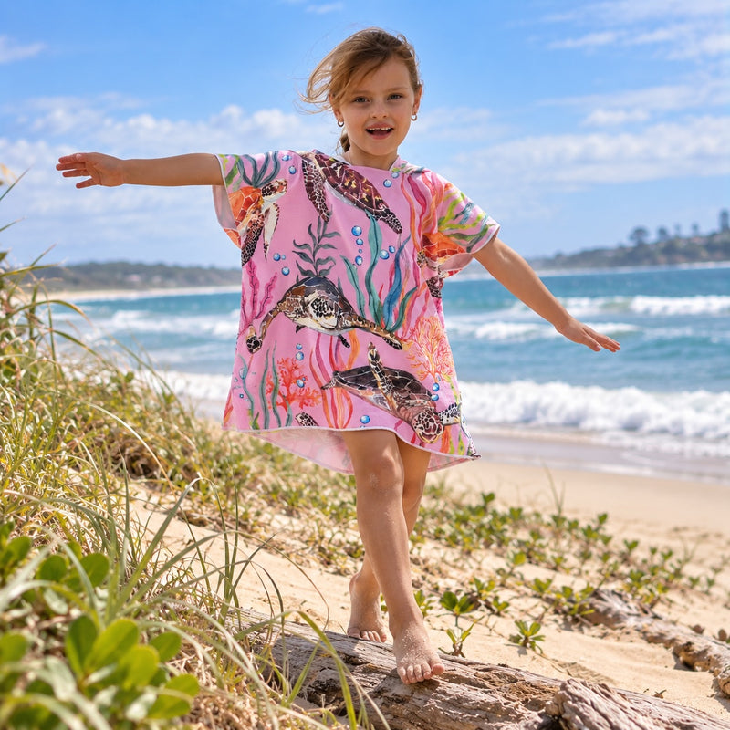 A young girl stands barefoot on driftwood on a sandy beach, wearing a pink short-sleeve sand free hoodie with a colorful underwater sea turtle print. The background shows ocean waves, blue sky, and beach grass.