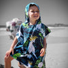 Boy (ages 6-12) wearing a blue hooded beach hoodie with a Bird of Paradise design featuring a yellow crested white cockatoo and palm leaves. He is standing at Currumbin Beach, Queensland, with one foot placed on a rock and the other in the shallow water. He has a relaxed and smile expression on his face.